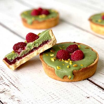 Фисташковый ганаш, малиновая прослойка в корзиночке из песочного теста. Сверху тарталетка украшена ягодами малины и дроблёной фисташкой.