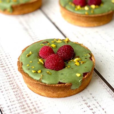 Фисташковый ганаш, малиновая прослойка в корзиночке из песочного теста. Сверху тарталетка украшена ягодами малины и дроблёной фисташкой.